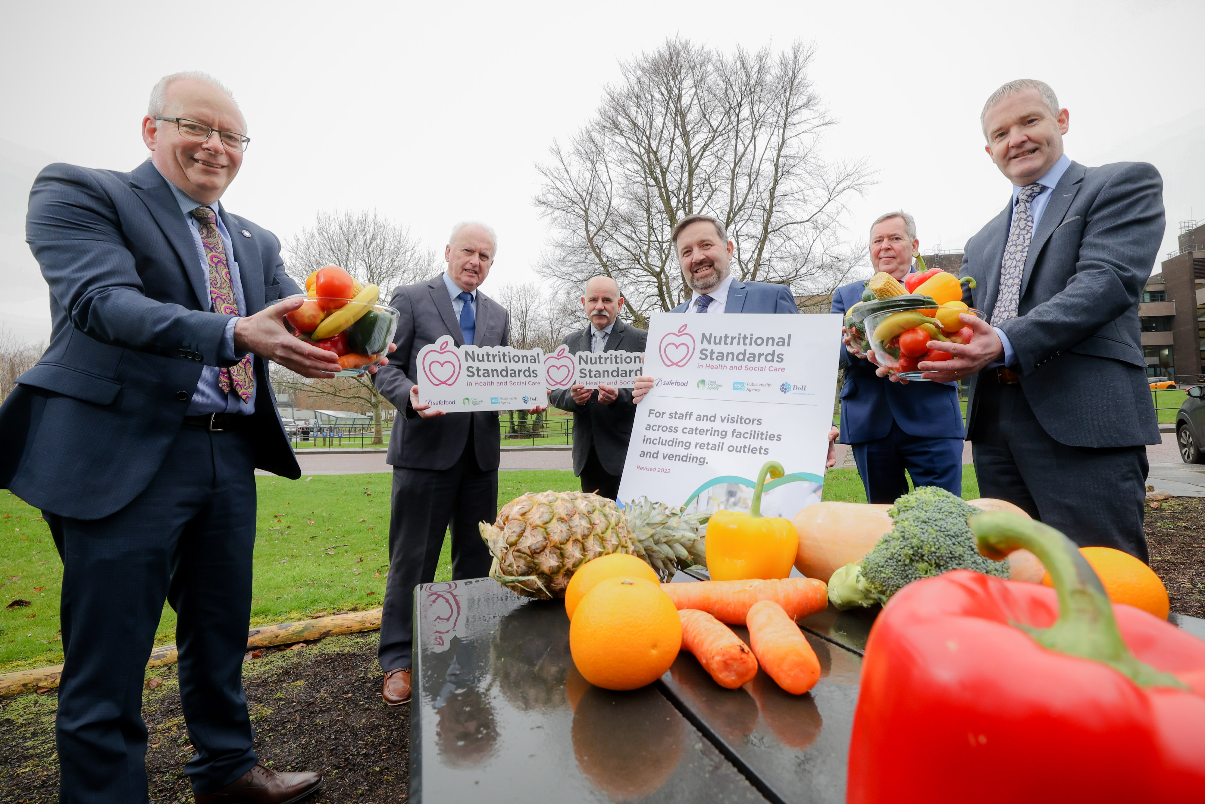 Six food and health officials show a report with a table of fresh fruit in the foreground